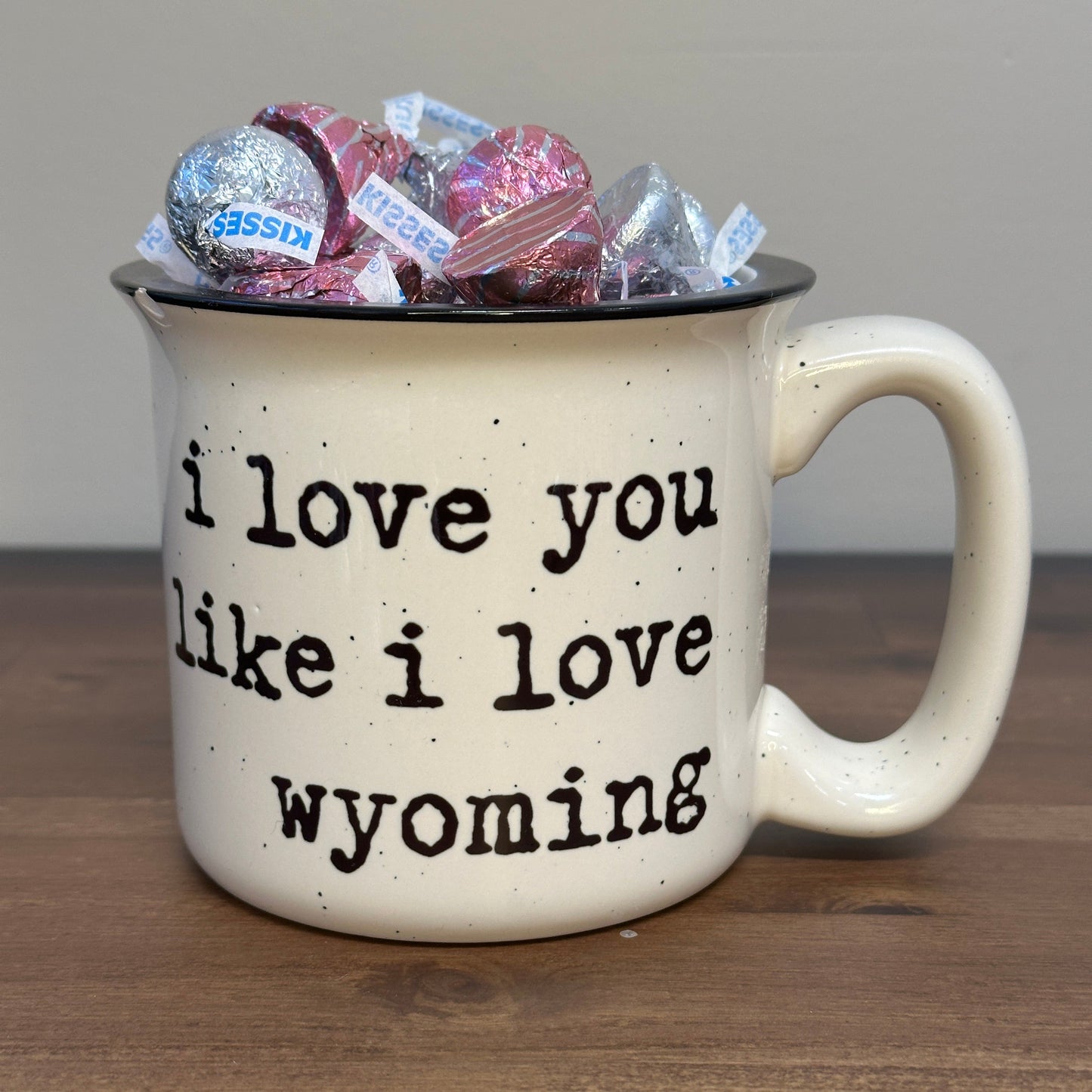 Cream mug with a thick handle and black colored rim that says 'I love you like I love Wyoming' text and candy inside on a wooden table.