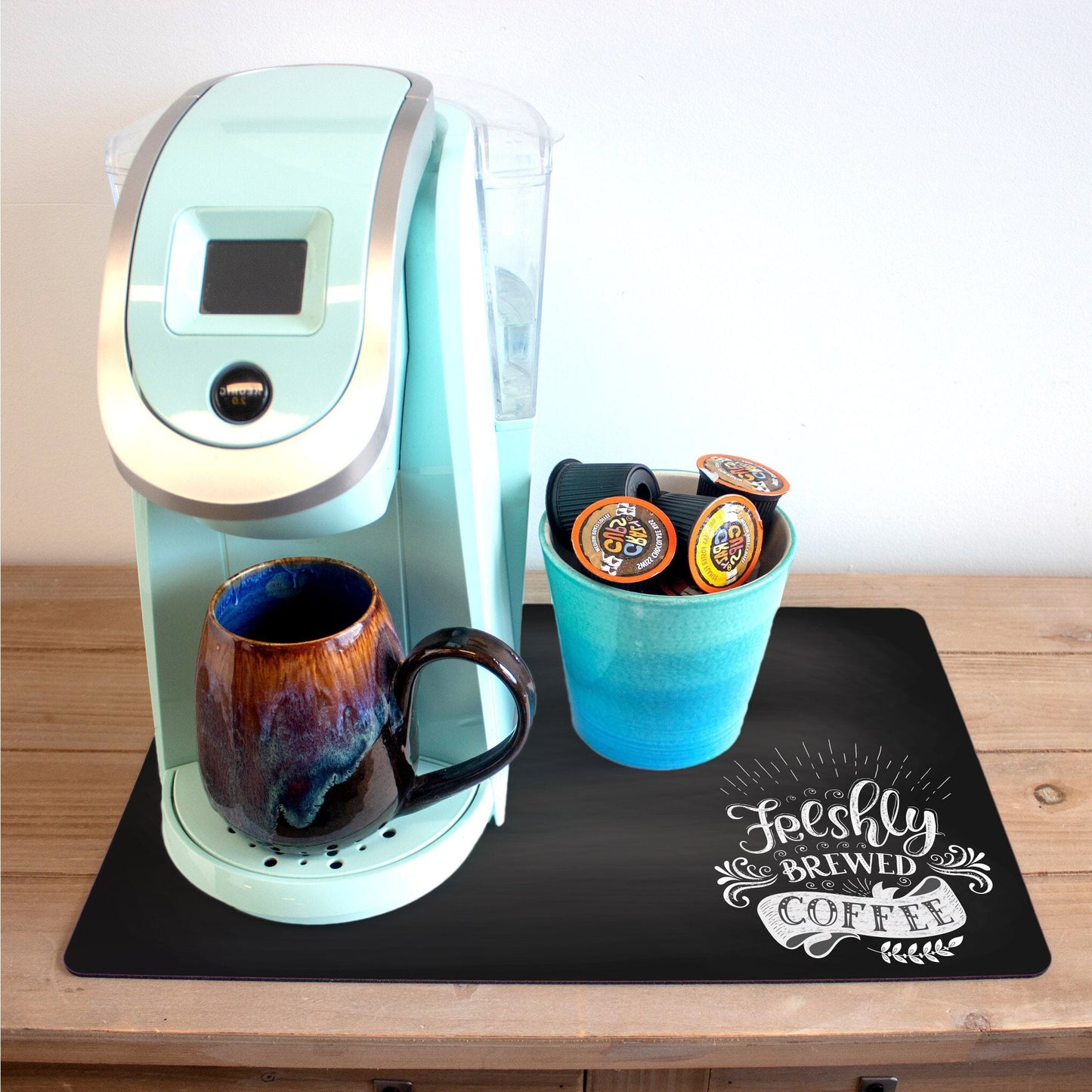 A light blue coffee maker sits on a wooden countertop, accompanied by a ceramic mug with a marbled blue and brown glaze and a turquoise cup holding several coffee pods. The setup is placed on a black coffee mat featuring the phrase "Freshly Brewed Coffee" in a whimsical white font.