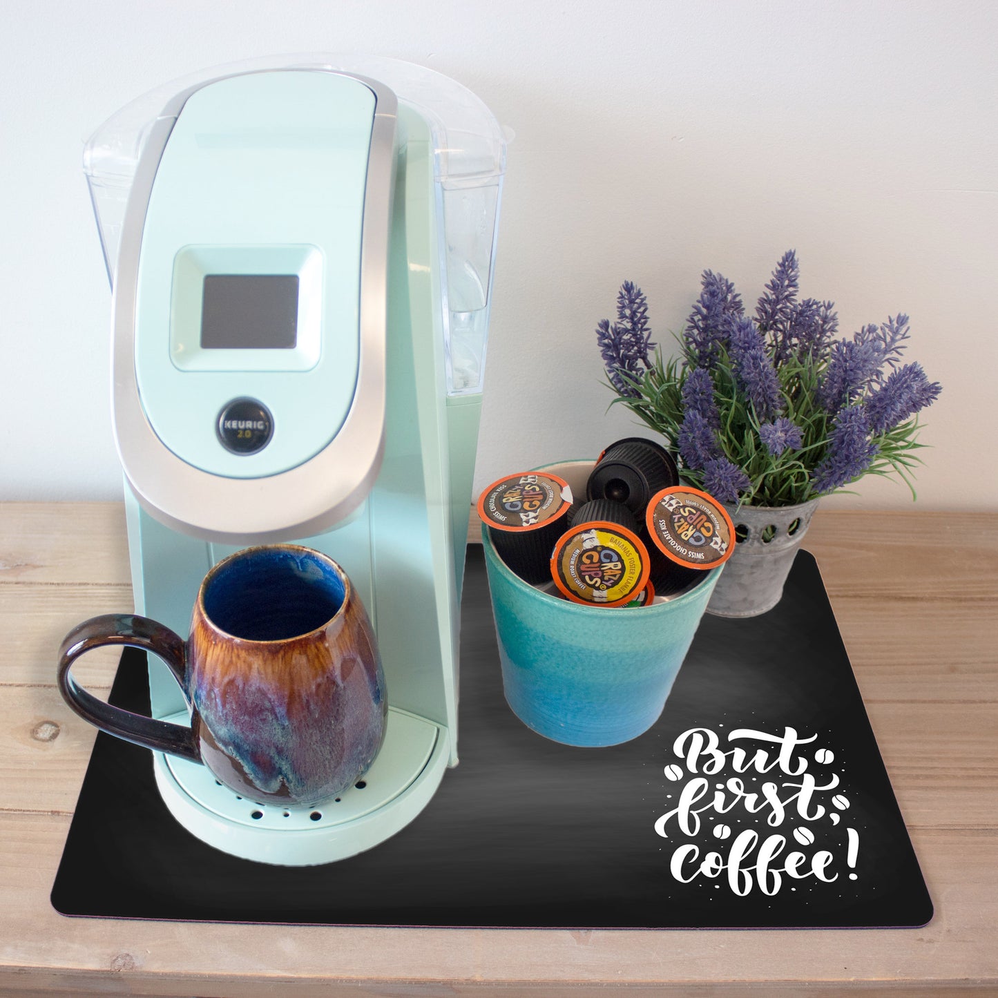 A light blue coffee maker on a wooden countertop with a ceramic mug that has a marbled blue and brown glaze and a turquoise cup holding several coffee pods. The setup is placed on a black coffee mat featuring the phrase "But First, Coffee!" in a whimsical white font. A small potted lavender plant is placed next to the coffee maker.