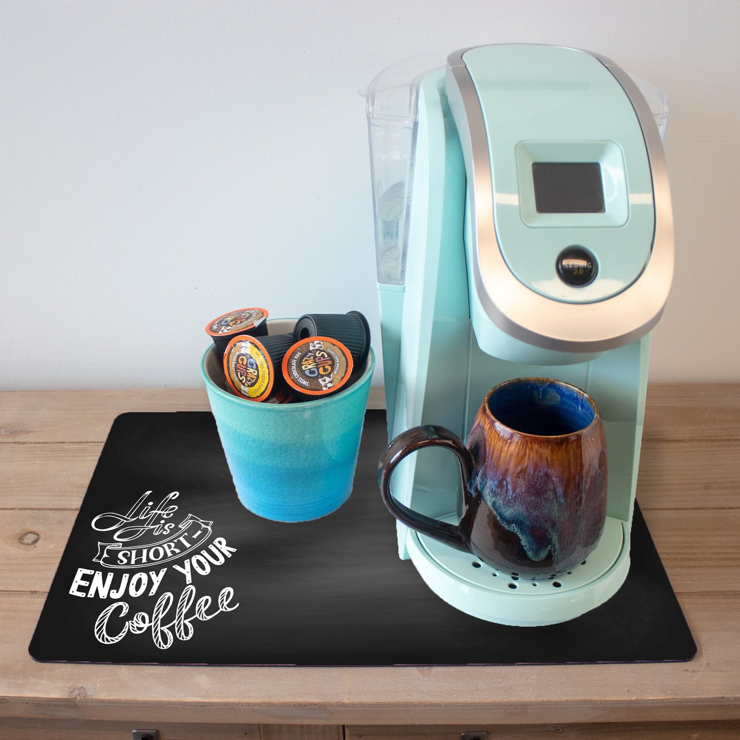 A light blue coffee maker on a wooden countertop with a ceramic mug that has a marbled blue and brown glaze and a turquoise cup holding several coffee pods. The setup is placed on a black coffee mat featuring the phrase "Life is Short, Enjoy Your Coffee" in a stylish white font.